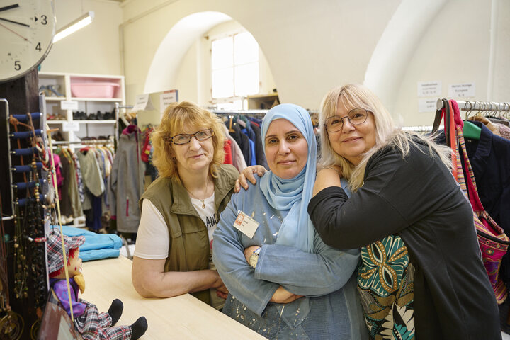 Drei Mitarbeiterinnen des Kleiderladens, eine mit grüner Weste, eine mit blauem Hijab und eine mit schwarzer Jacke, lehnen lächelnd vor Kleiderständern und Regalen am Verkaufstresen.