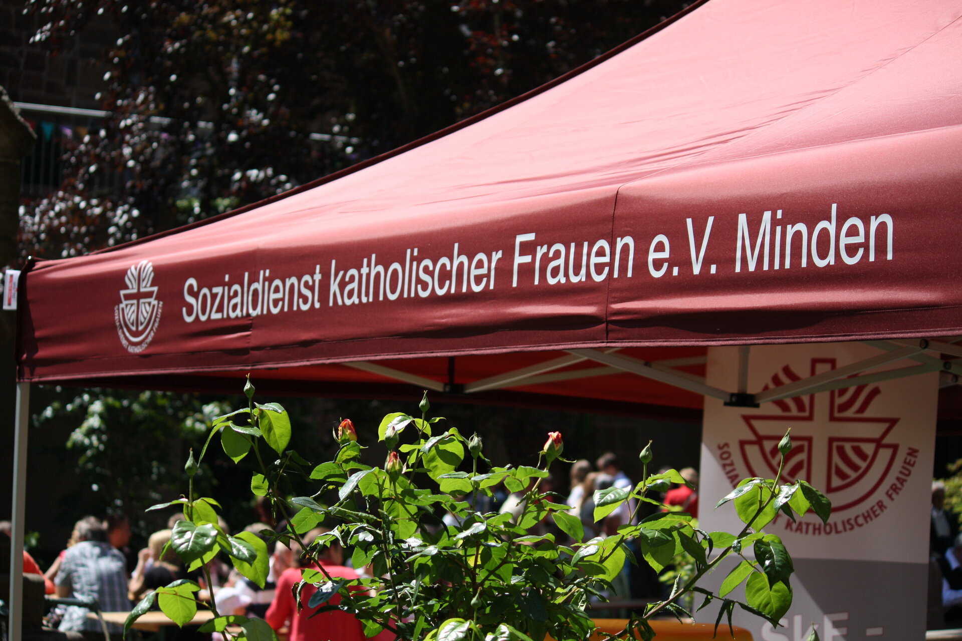 Ein rotes Zelt mit weißer Aufschrift "Sozialdienst katholischer Frauen e. V. Minden" steht in einem Park, begleitet von einer weißen Fahne und einem Baum mit roten Blumen.